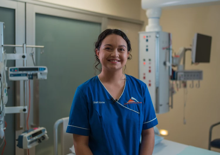 A staff nurse standing in a hospital room in front of a vacant hospital bed smiling