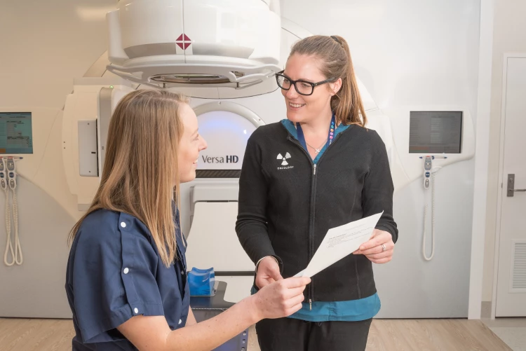 A Radiation Therapist hands out some aftercare paperwork to a patient. Behind them is a Versa HD machine.